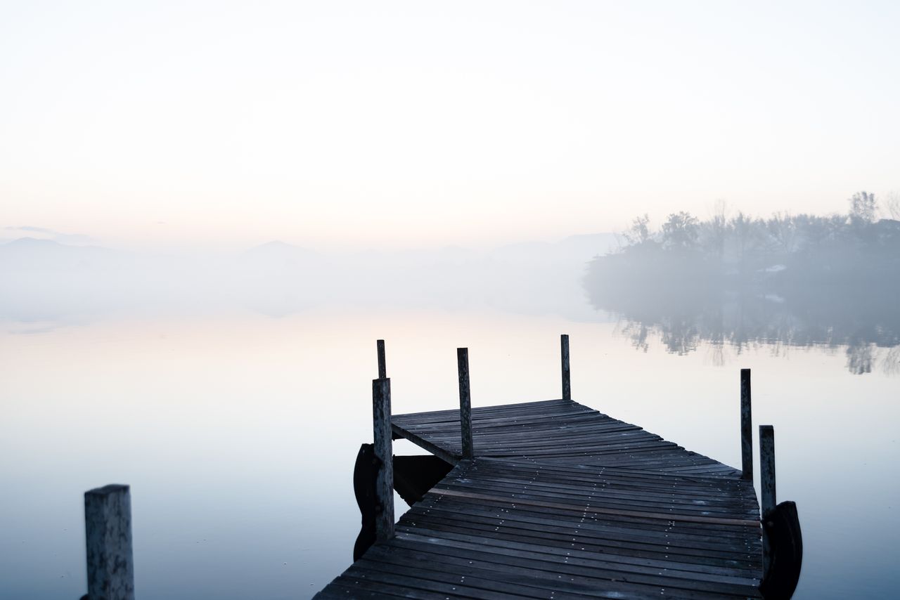 Home A tranquil lakeside view at dawn with mist enveloping the boardwalk and distant trees.