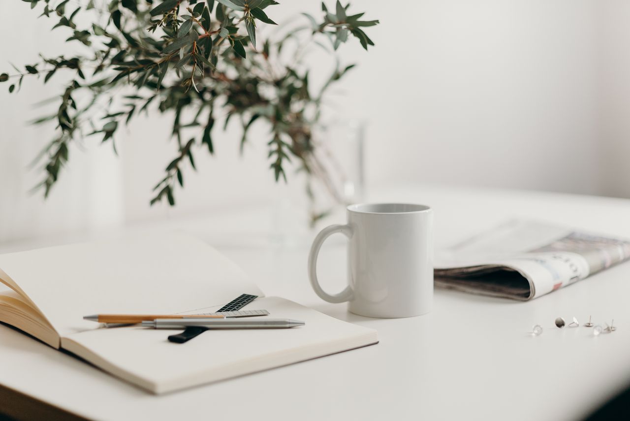 Home A serene minimalist workspace featuring a white coffee mug, open notebook, and green plant vase on a desk.