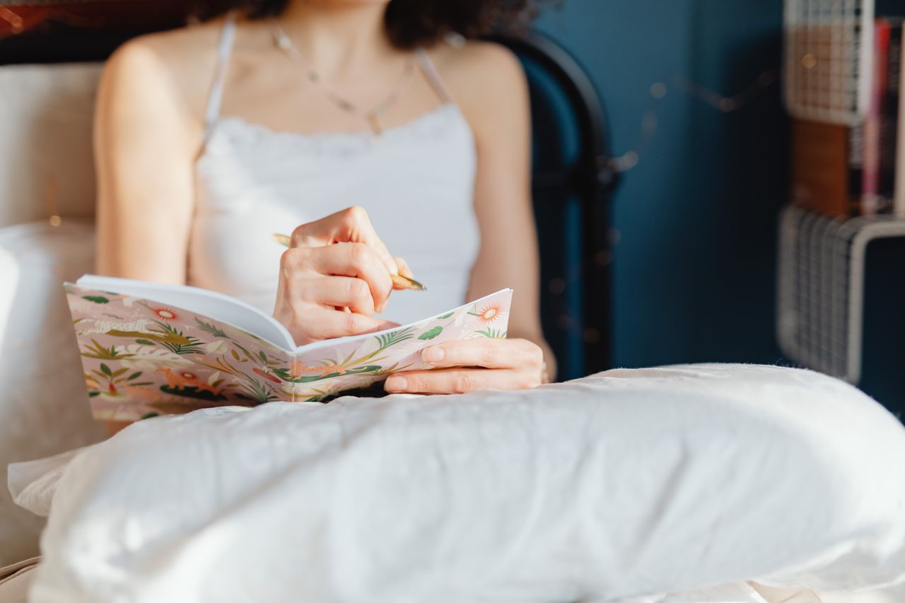 Home A woman in a white top writes in a decorative floral notebook while seated on her bed.